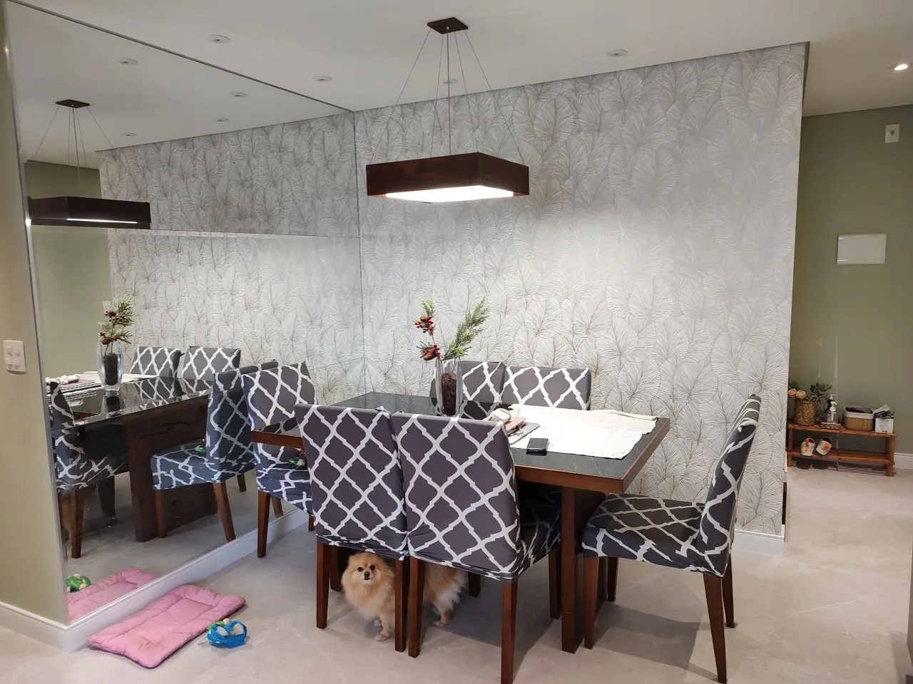 Sala de jantar com mesa de madeira escura, cadeiras de estampa geométrica, parede espelhada e papel de parede de folhagens; um cachorro sob a mesa.