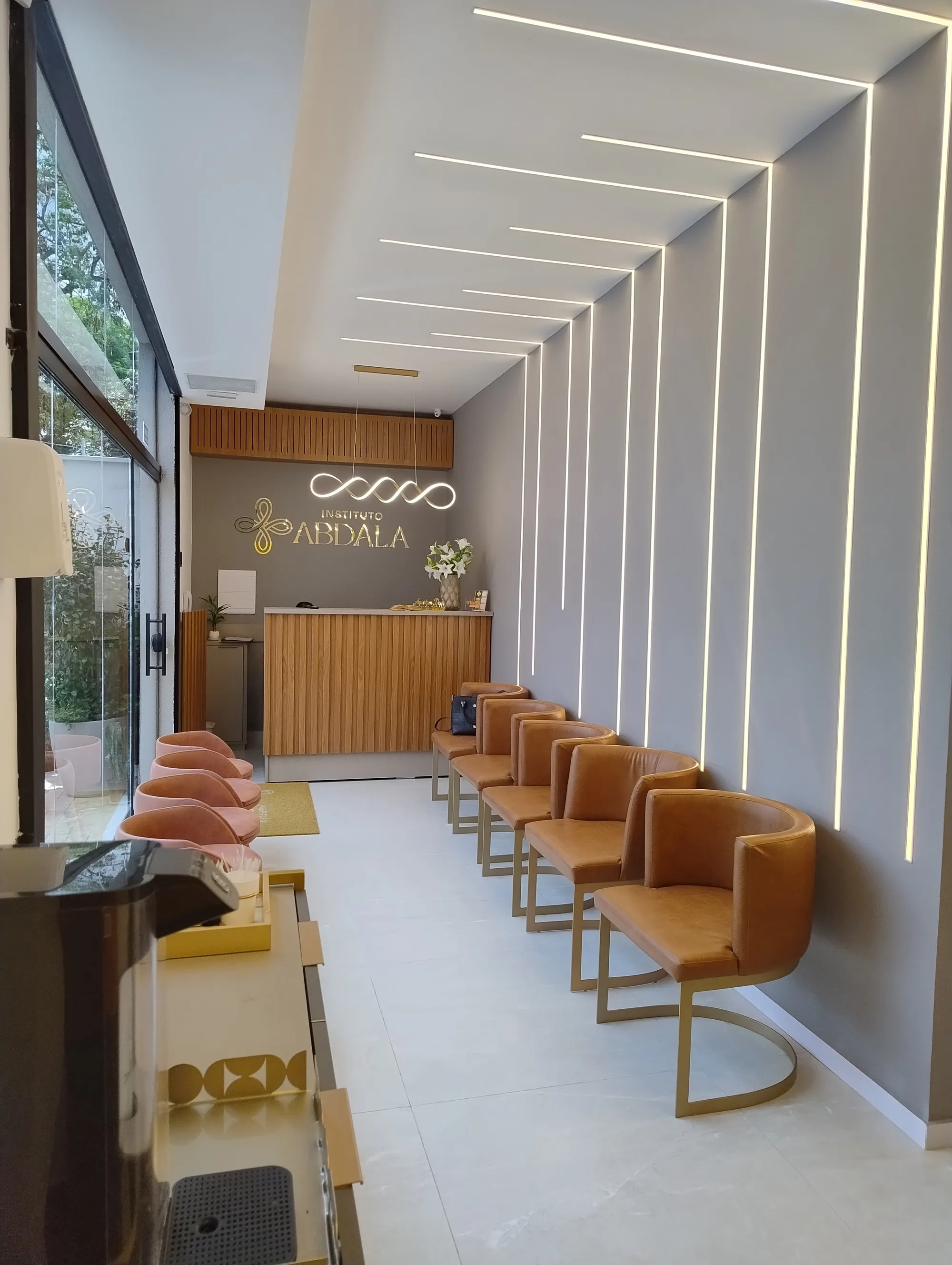 Recepção com balcão em madeira ripada, poltronas em couro caramelo, pendente moderno em LED e iluminação linear na parede e teto.