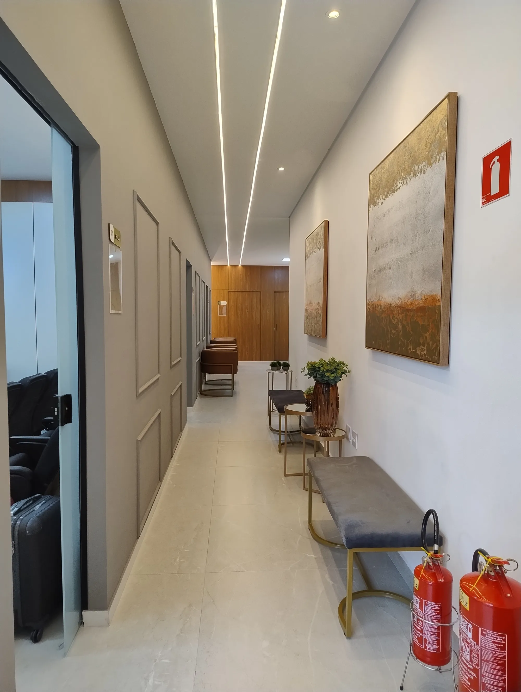 Corredor com paredes cinzas e molduras tipo boiserie, piso claro, quadros abstratos, banco de veludo e perfis de iluminação LED contínuos.