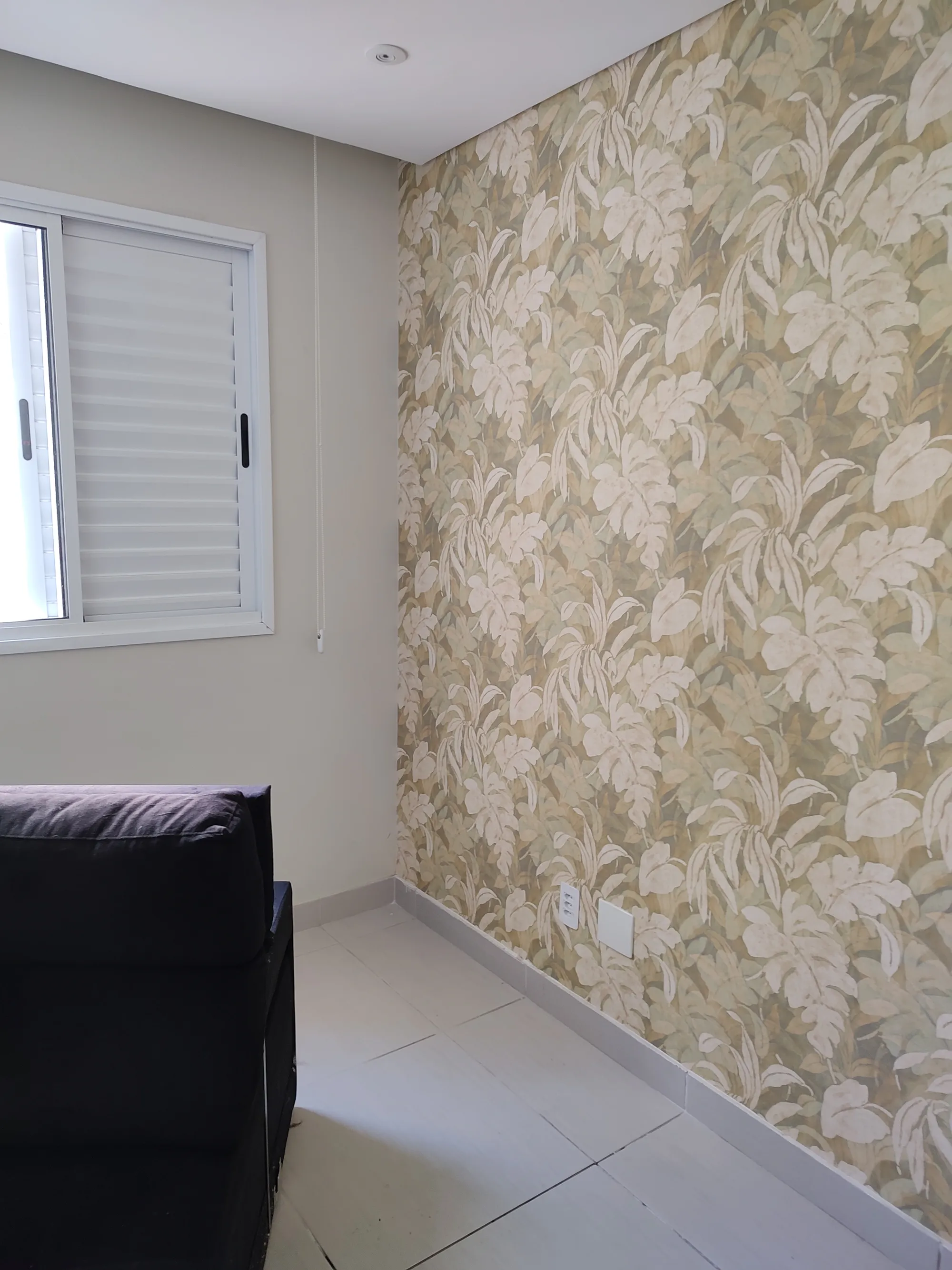 Canto de um ambiente com detalhe de um sofá escuro e parede em destaque revestida com papel de parede de estampa tropical em tons de verde e bege.