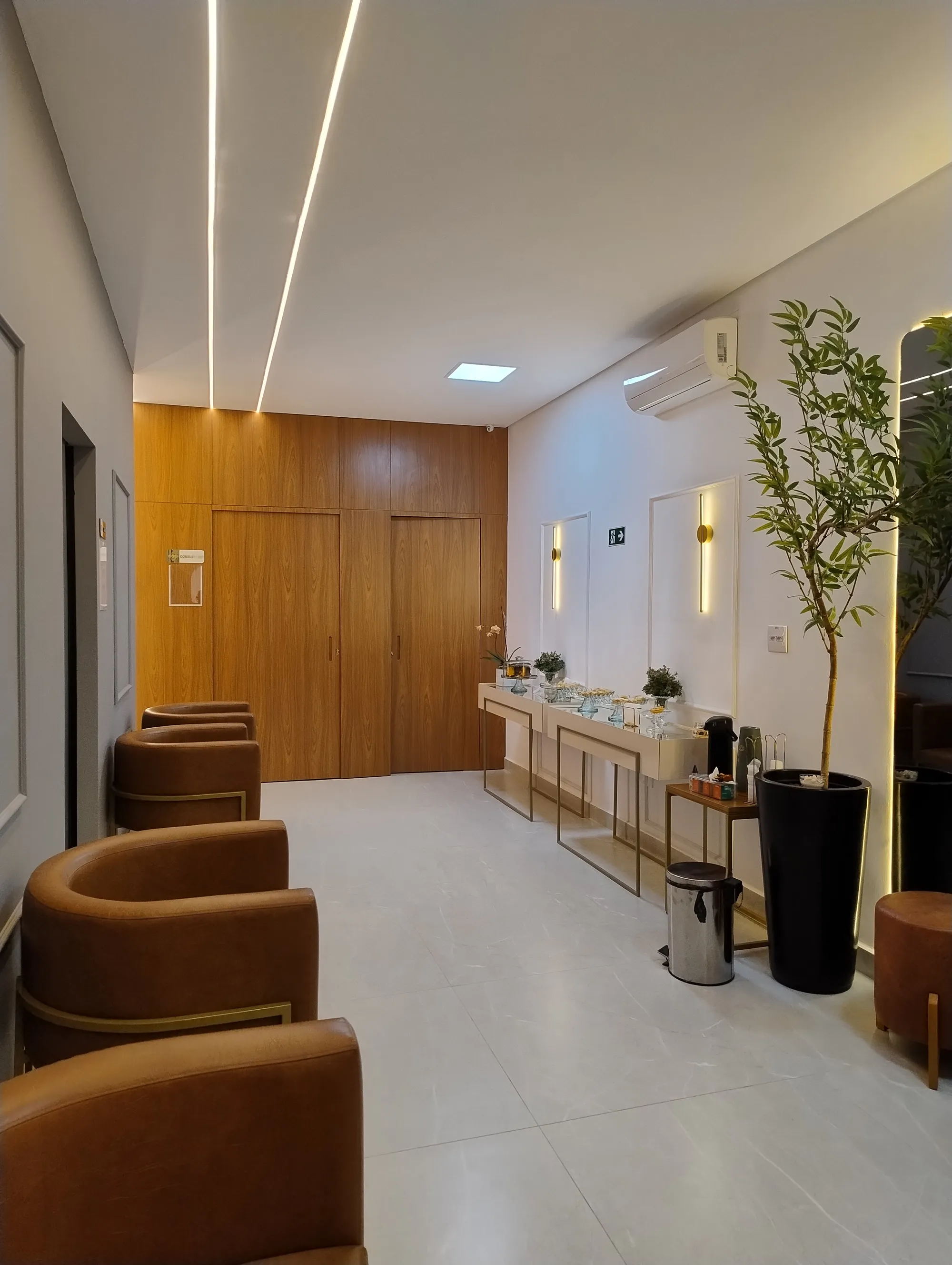 Área de espera com poltronas caramelo, portas em painel de madeira, espelho iluminado e aparador com itens de café.