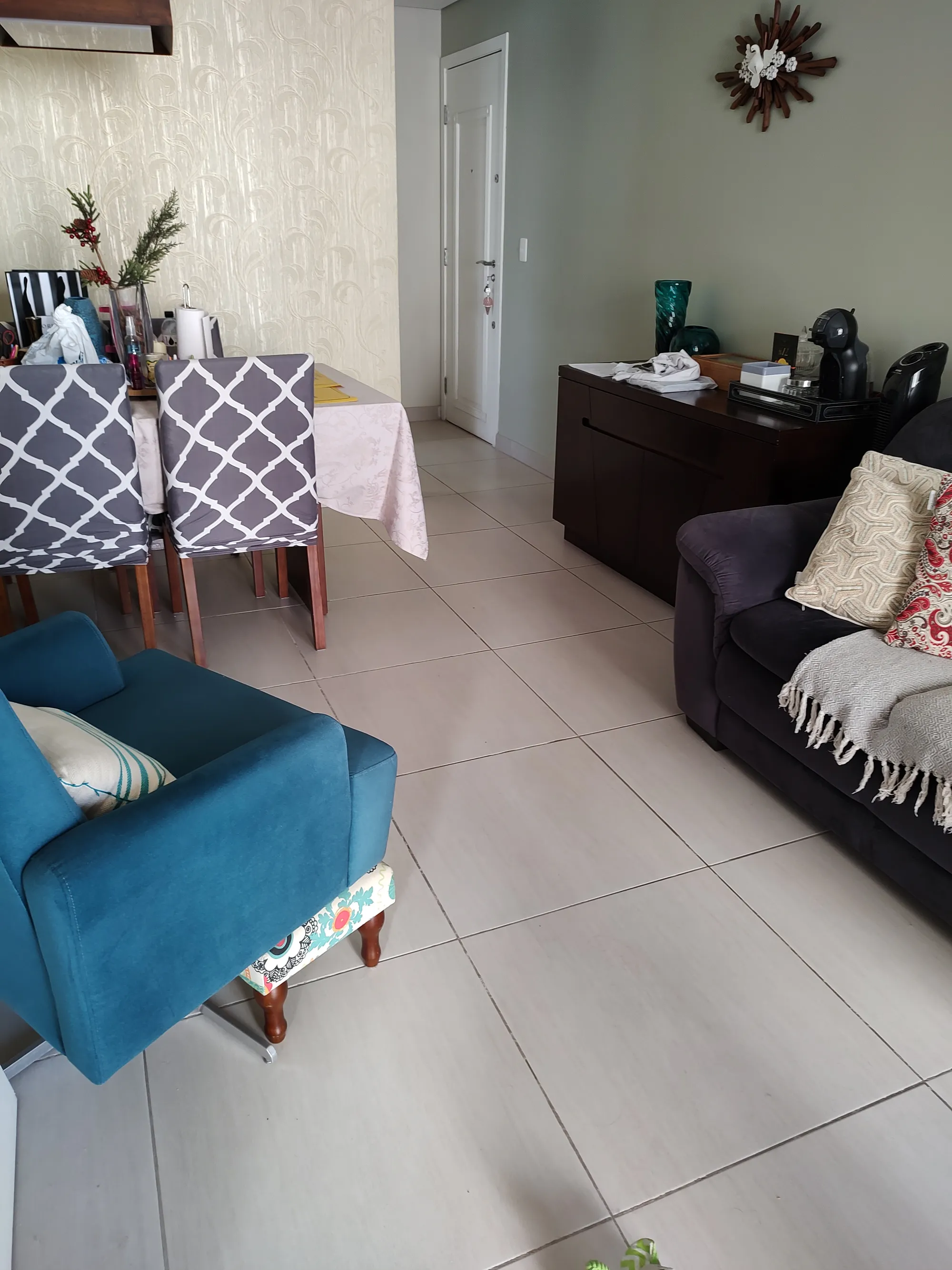 Sala de estar com poltrona azul turquesa, sofá escuro com almofadas, aparador de madeira com cafeteira e vista parcial da sala de jantar integrada.