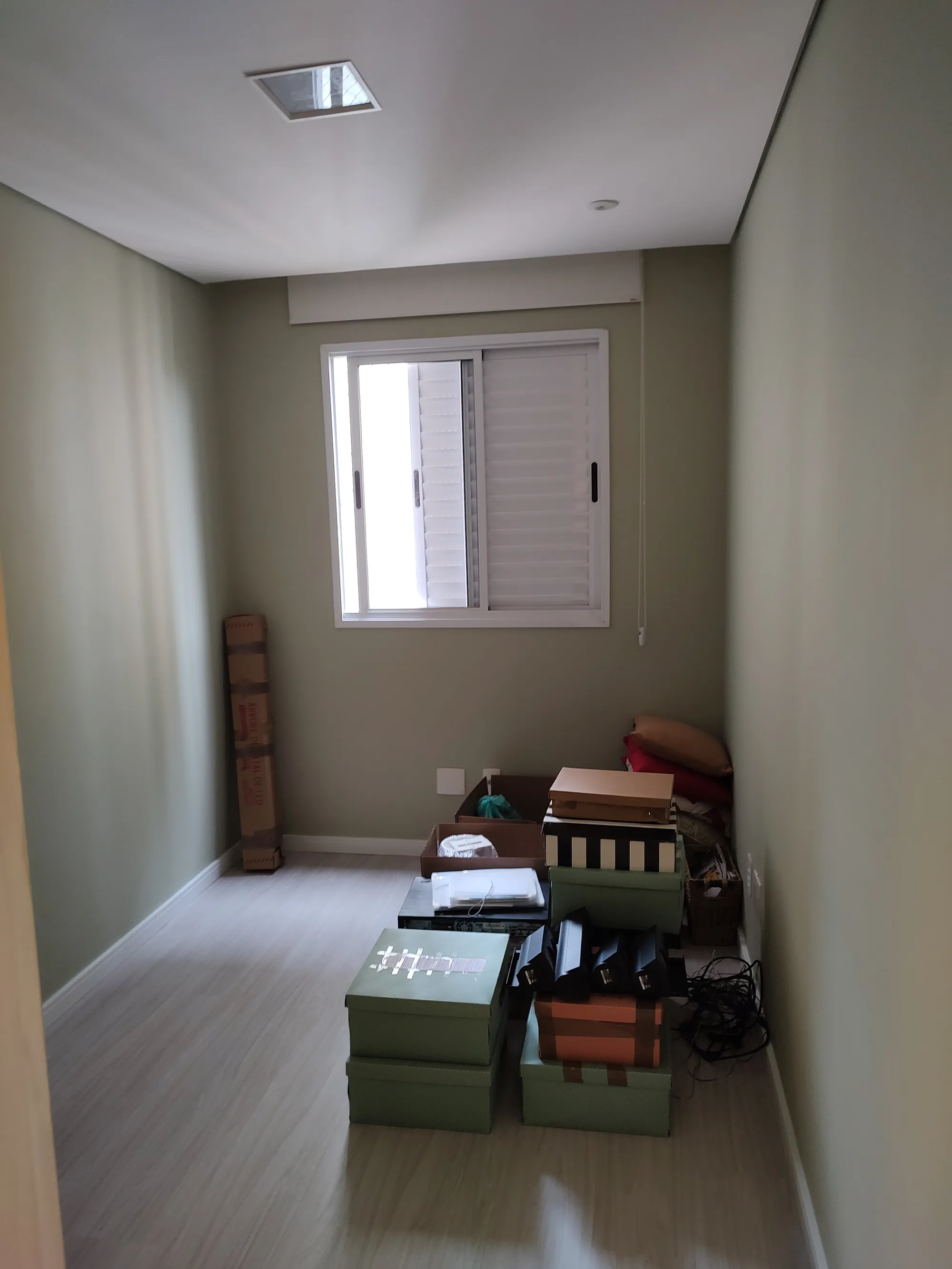 Quarto com piso laminado claro, paredes verde-claras, janela branca e diversas caixas de papelão empilhadas no chão.