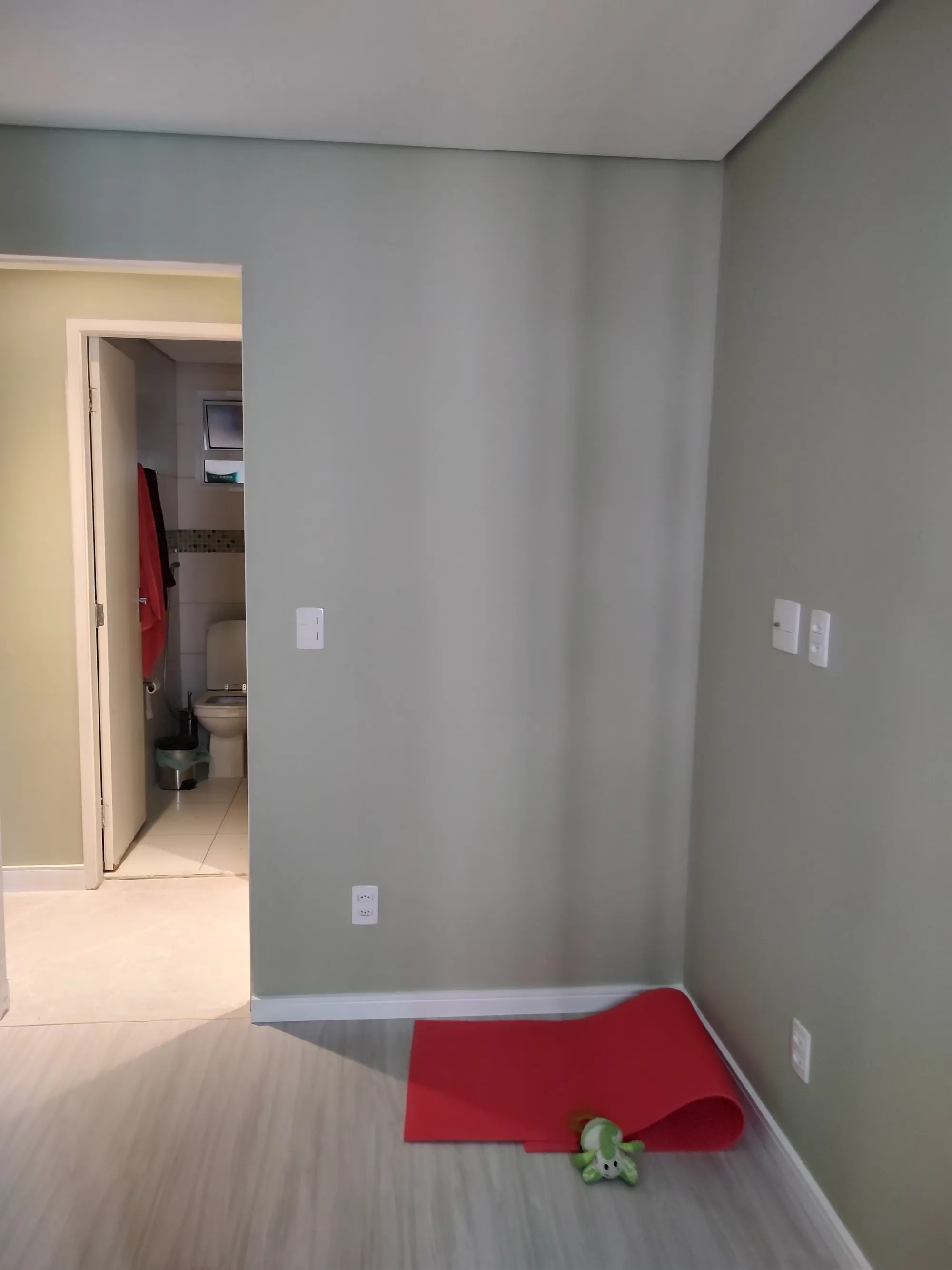 Quarto com piso laminado, paredes verde-claras, um tapete de yoga vermelho estendido no chão e vista para a porta aberta de um banheiro claro.