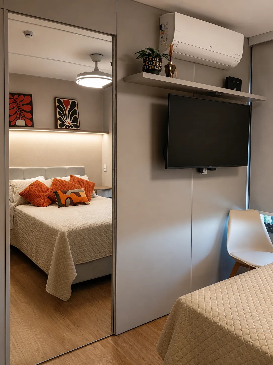 Visão do quarto mostrando um guarda-roupa com porta de espelho, painel de parede cinza com TV instalada, ar-condicionado e parte da cama com almofadas laranjas.