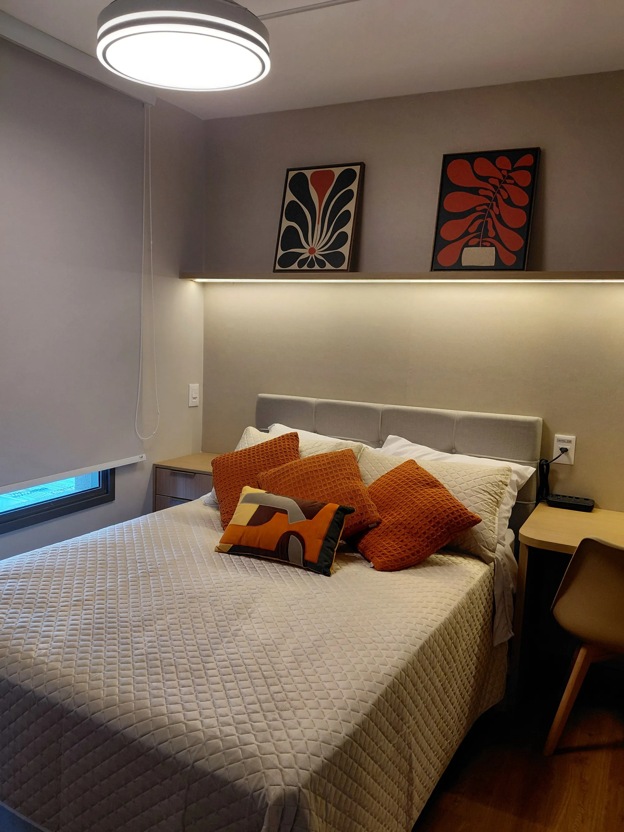 Cama de casal com cabeceira estofada cinza e almofadas laranja; acima, uma prateleira com fita de LED ilumina dois quadros decorativos.