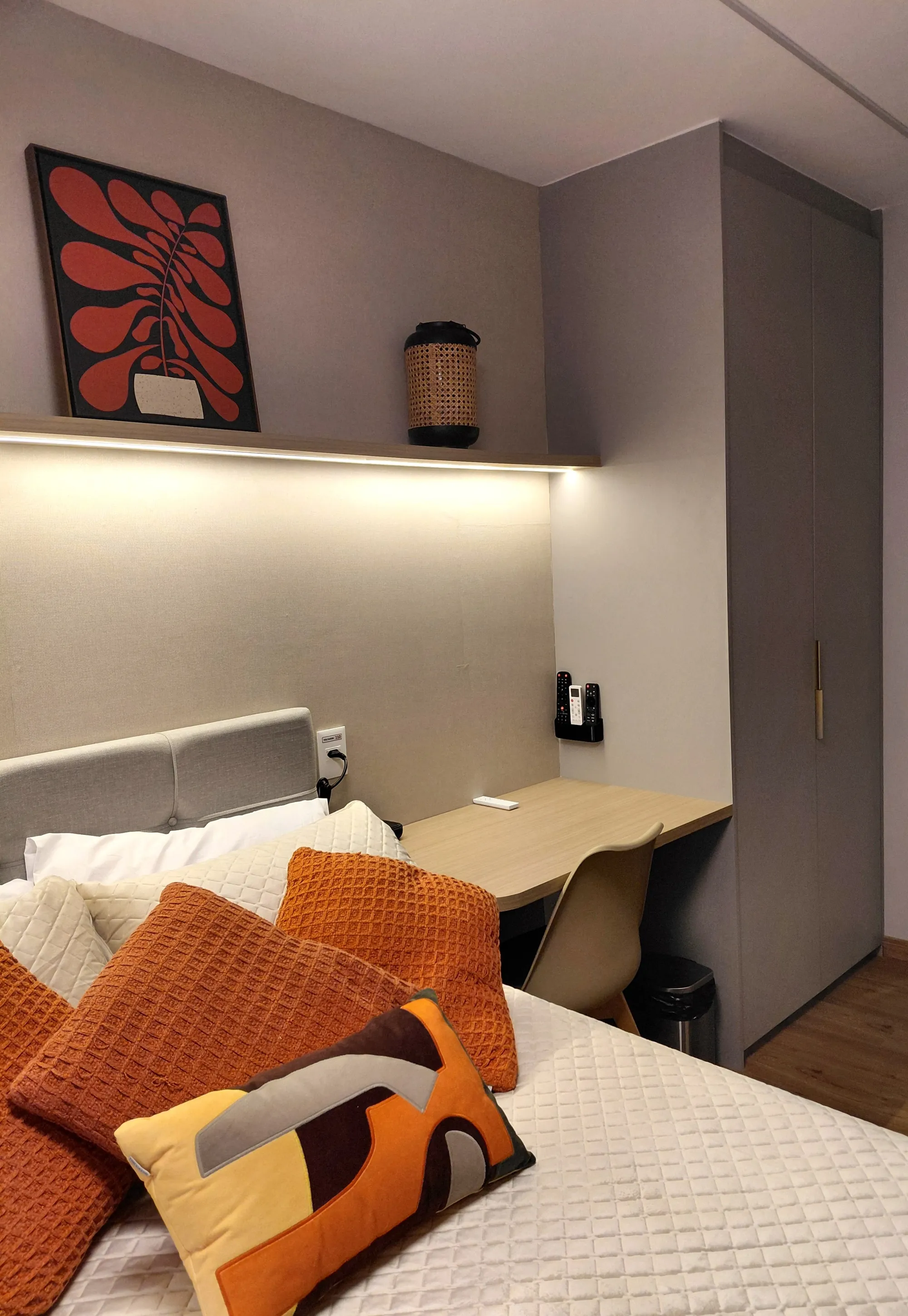 Detalhe em ângulo da cama com almofadas coloridas, mostrando a bancada de trabalho ao fundo, a iluminação quente da prateleira e a lateral do armário cinza.